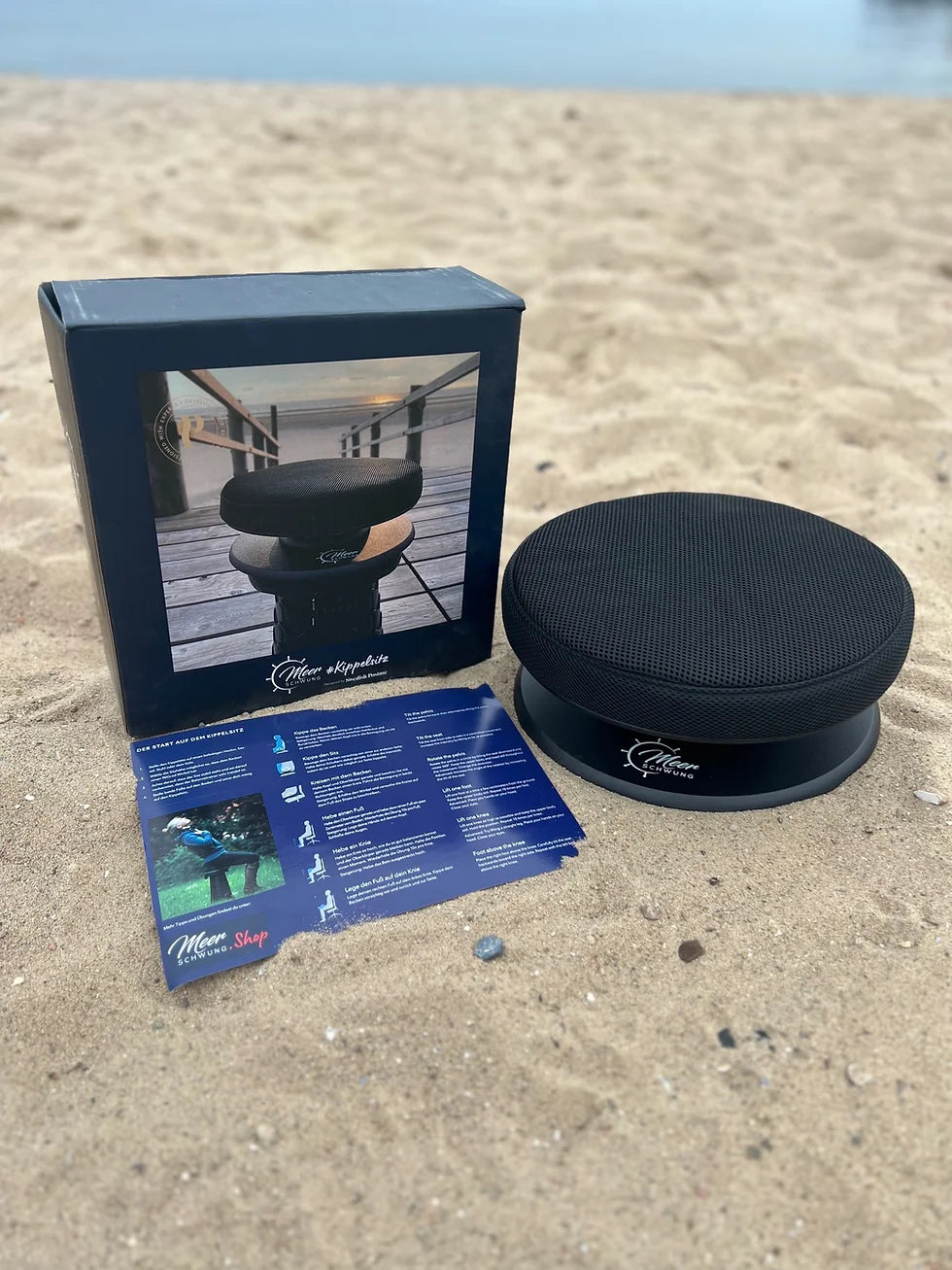Ergonomischer Kippelsitz zur Aktivierung der Core-Muskulatur für Reiter - platziert am Sandstrand neben Produktkarton und beiliegender Anleitung